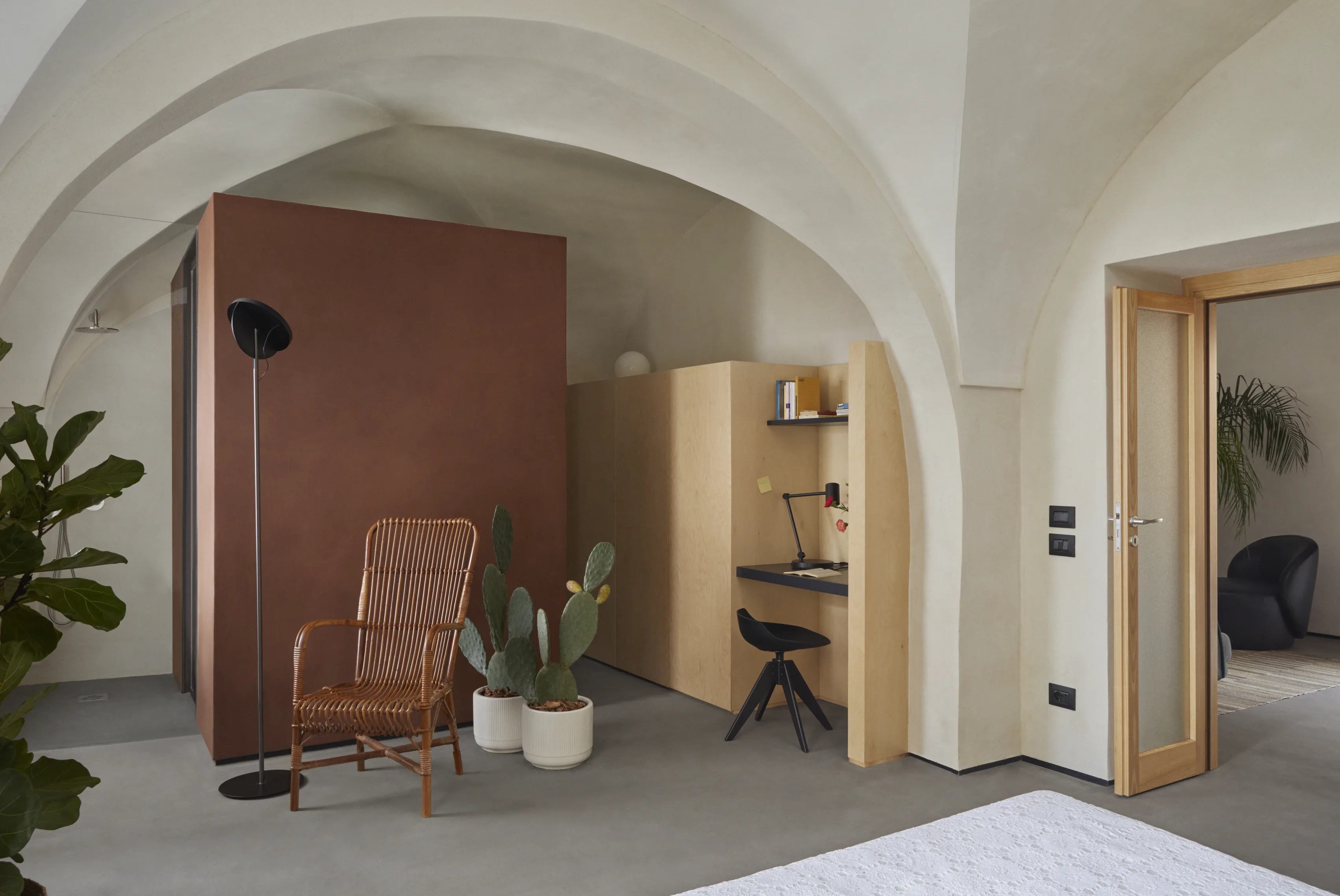 Bedroom with white cross vaults, a corten steel cube, a wicker armchair and a small desk