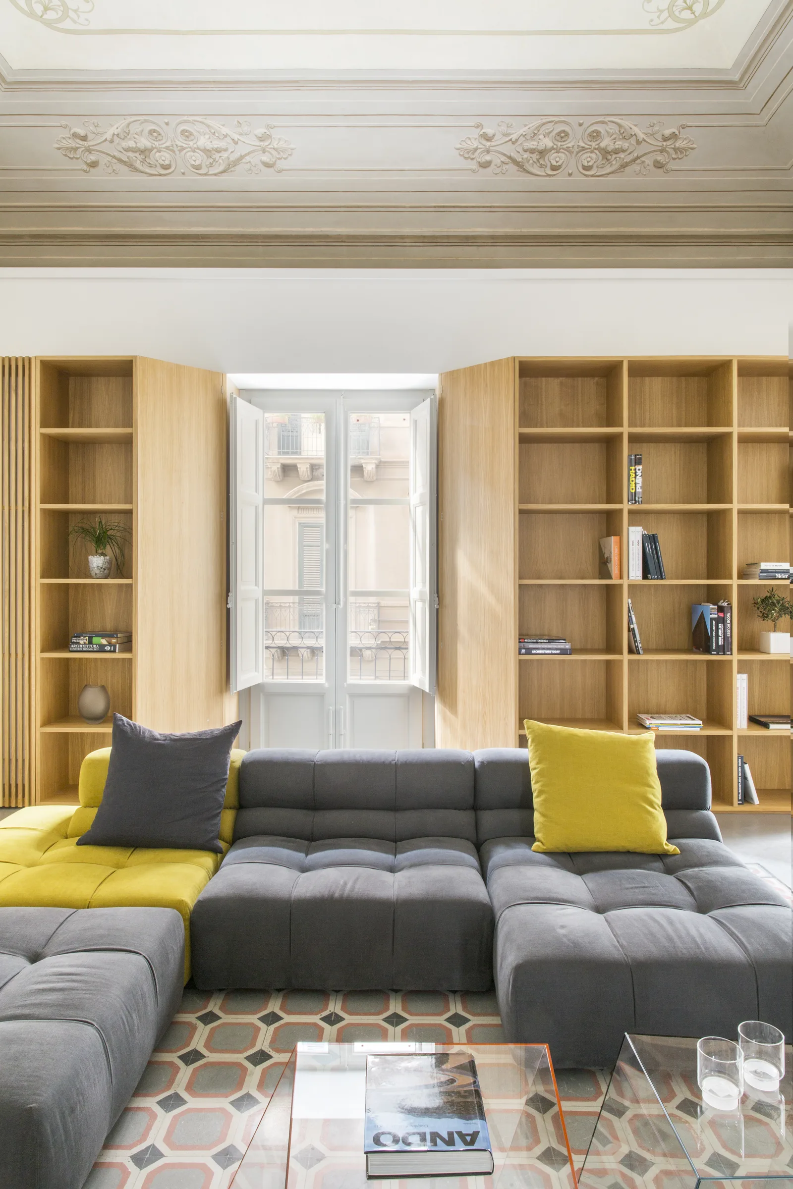 Grey and yellow sofa between two bookcases, in front of a window with closed shutters