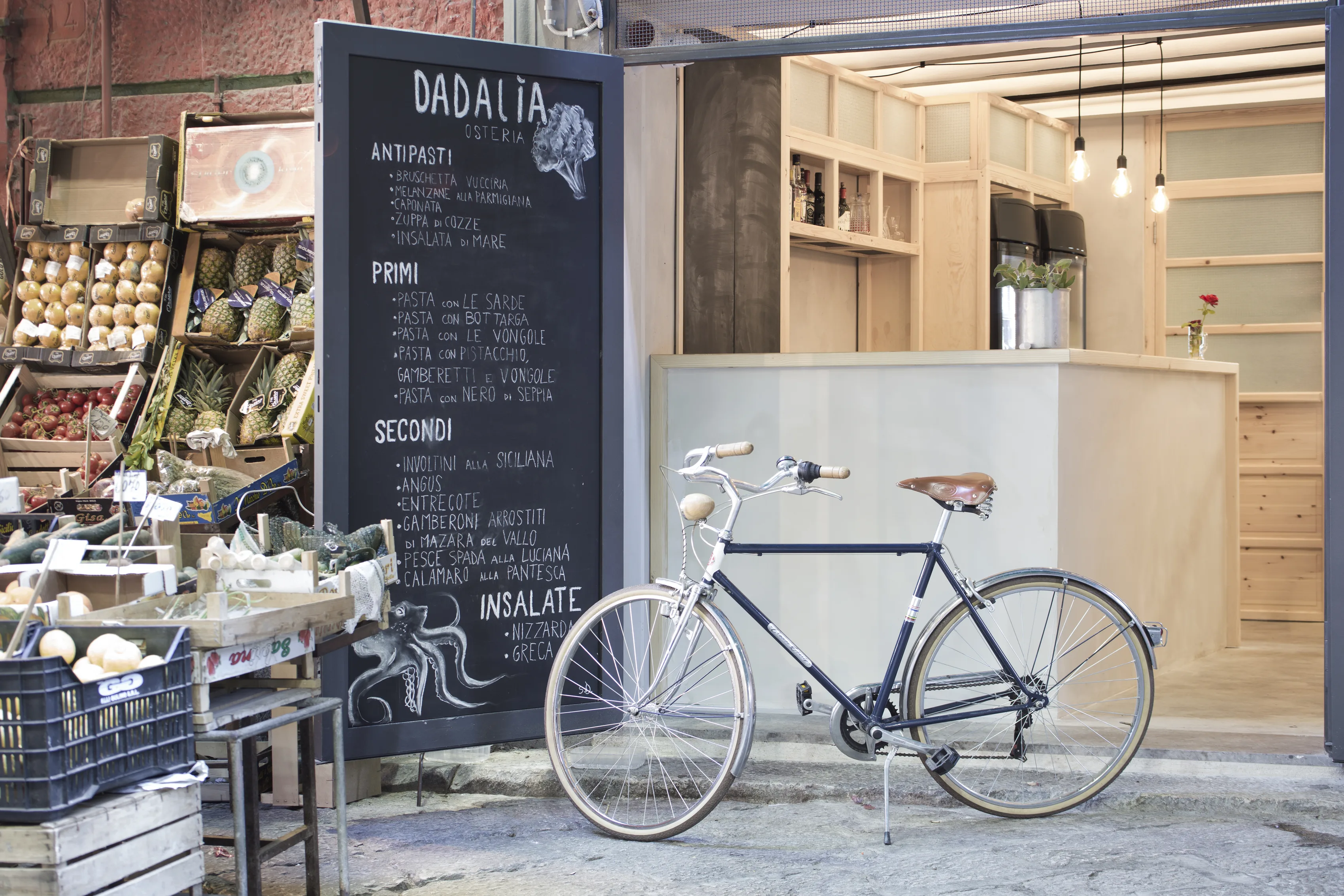 Ingresso dell'osteria su strada, con lavagna del menu, bicicletta e banco in legno chiaro