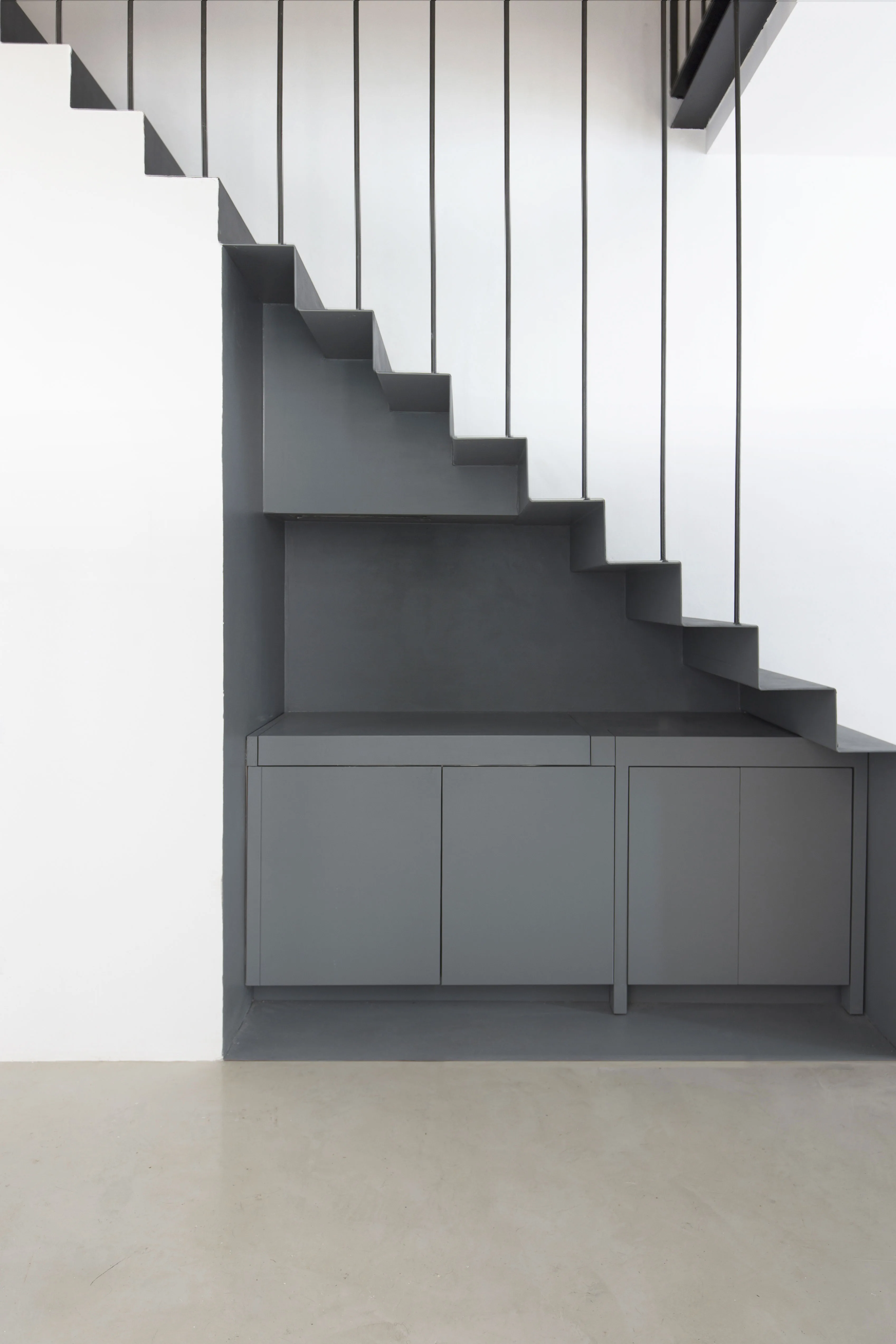 Stair with slender metal uprights and, beneath it, a grey compact kitchen with a mirrored splashback