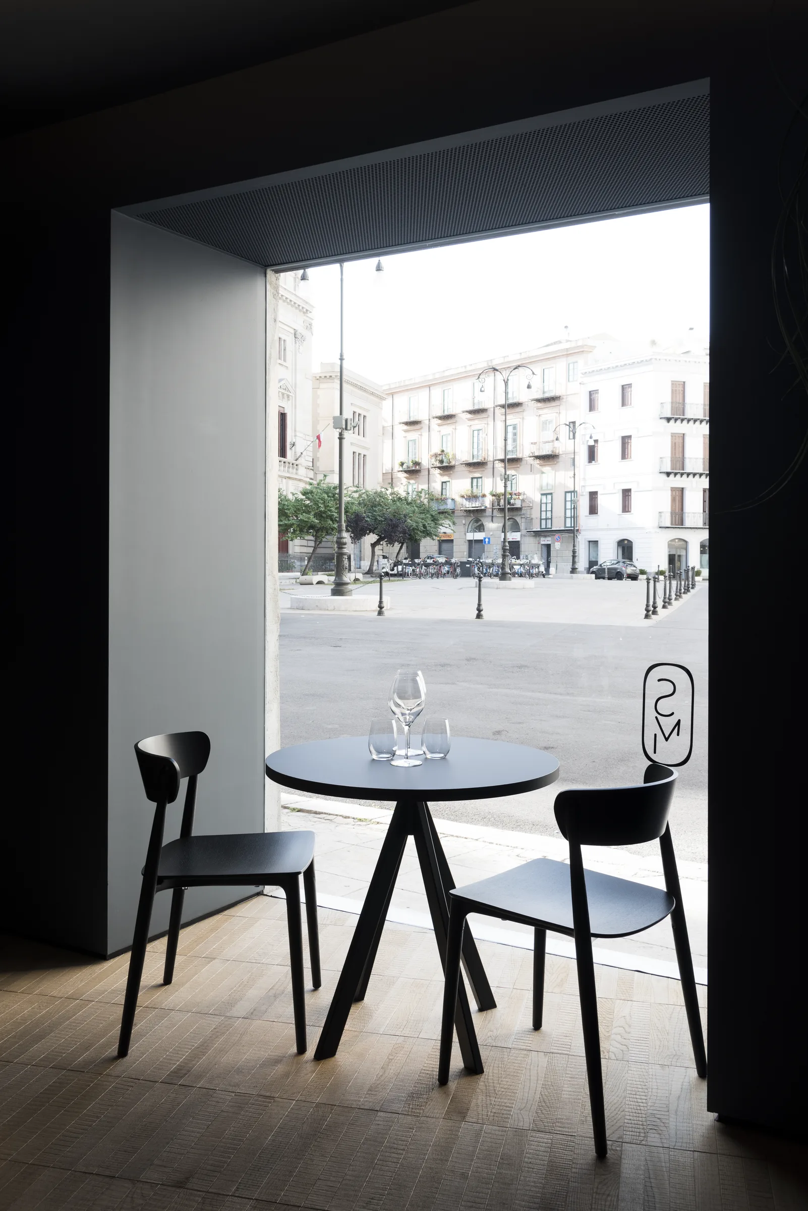 Tavolo tondo e due sedie nere davanti alla vetrata, con gli edifici della piazza fuori