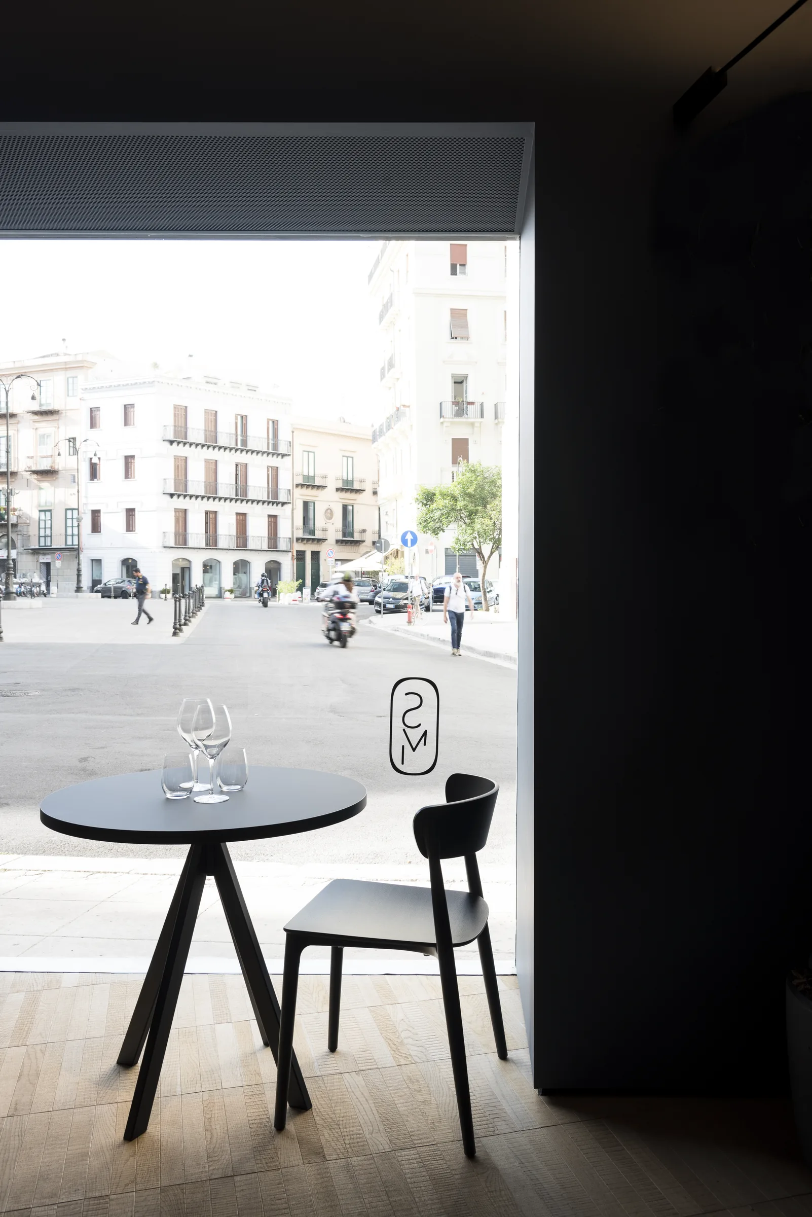 Tavolo con un bicchiere e sedie nere contro la luce della vetrata, inquadratura più ampia sulla piazza