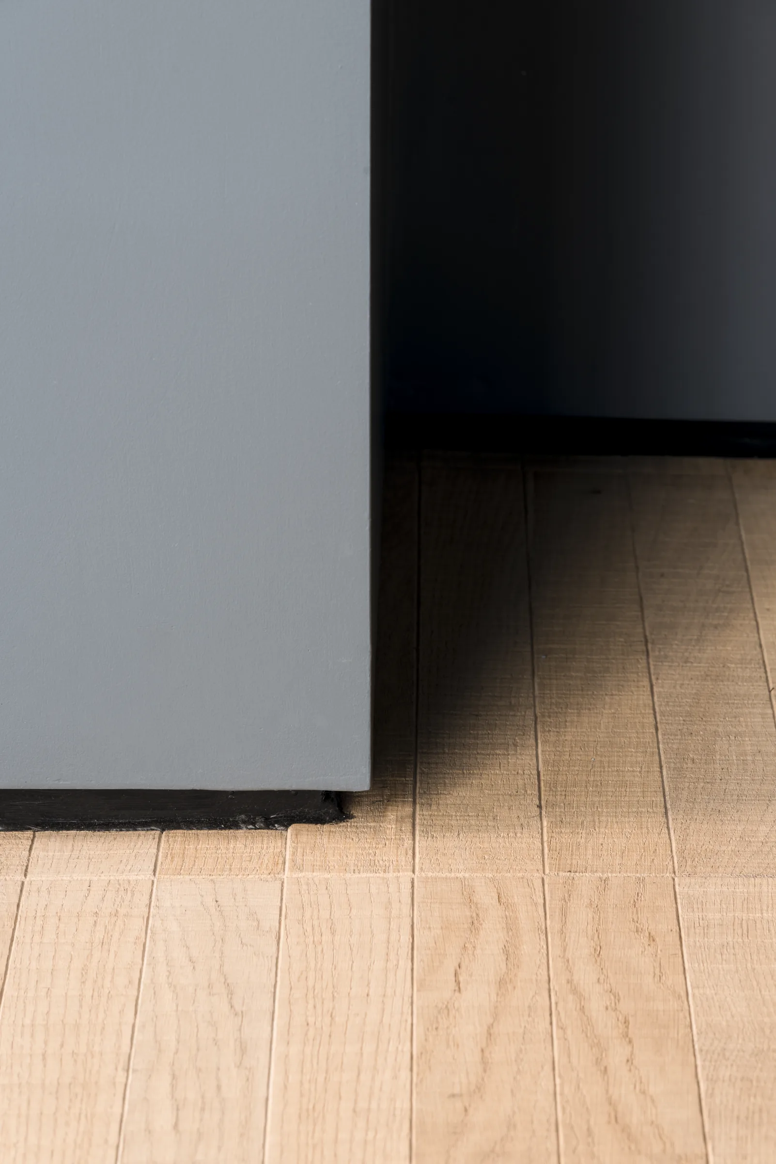 Parete grigio-azzurra che incontra il parquet a listoni, con ombra sul fondo