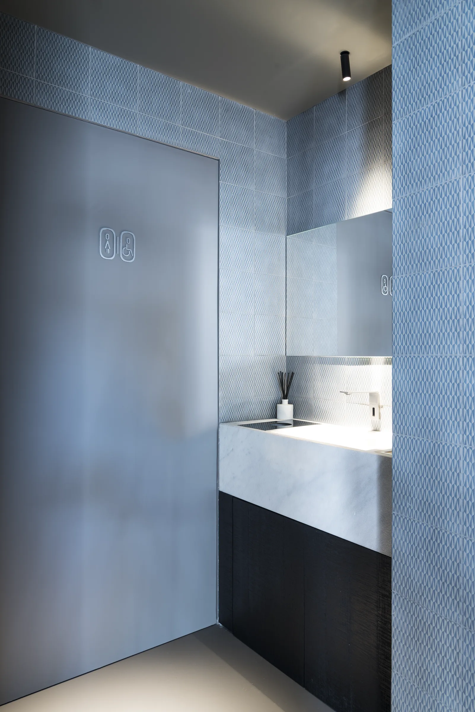 Bagno con mosaico azzurro, mensola lavabo in pietra chiara e faretto a soffitto, visto di lato