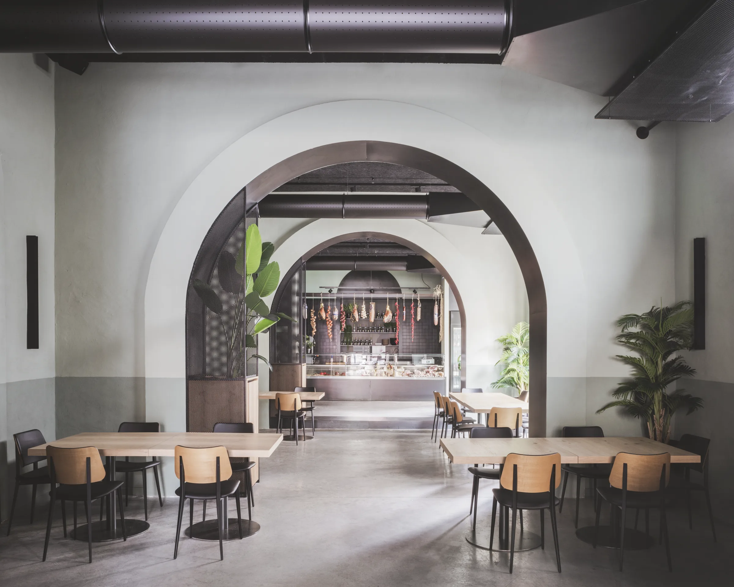 Sala con tavoli in rovere e sedie nere, e un doppio arco che inquadra il banco dei salumi sul fondo